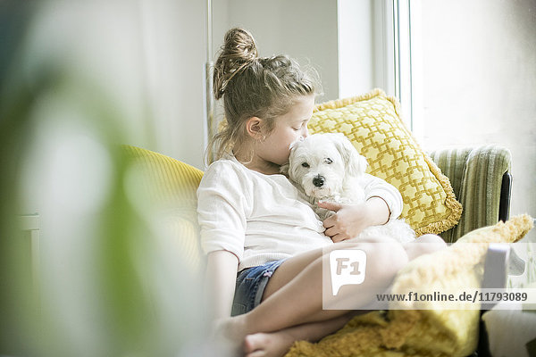 Little girl sitting on armchair at home cuddling with her dog