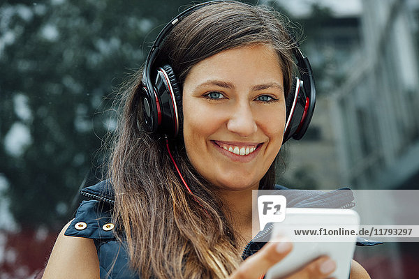Porträt der lächelnden jungen Frau mit Kopfhörer und Handy in der Stadt