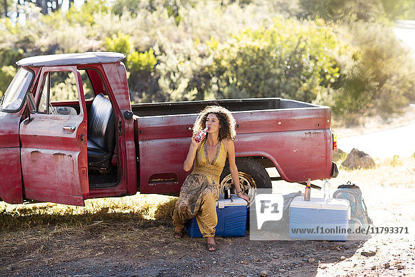 Junge Frau sitzt neben dem alten rostigen Pick-up und trinkt aus der Flasche.