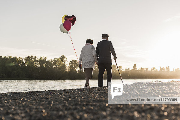 Rückansicht des Seniorenpaares mit Ballons die abends am Flussufer spazieren gehen.