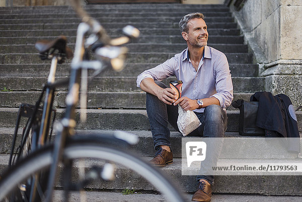 Lächelnder Mann sitzt auf einer Treppe mit Kaffee zum Mitnehmen in der Stadt.