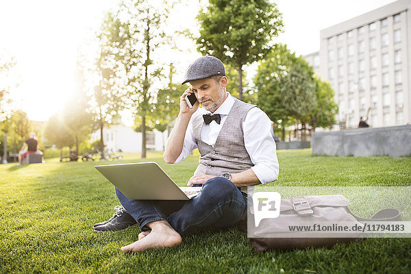 Confident mature businessman with laptop and smartphone in the city park sitting on grass