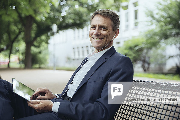 Smiling mature businessman sitting on park bench with tablet and earphones