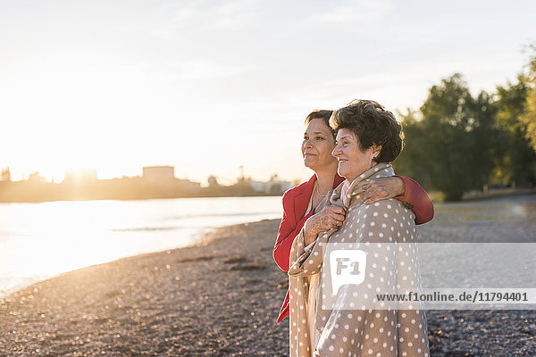 Fröhliche Seniorin mit ihrer erwachsenen Tochter am Strand bei Sonnenuntergang