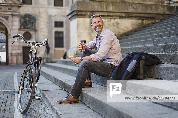 Lächelnder Mann sitzt auf einer Treppe mit Kaffee zum Mitnehmen in der Stadt.