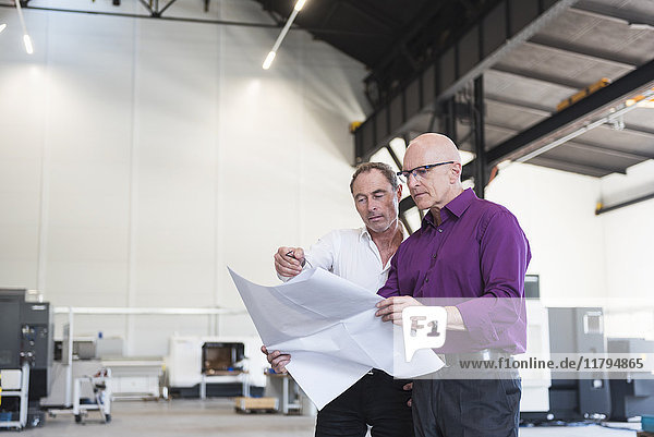 Zwei Geschäftsleute schauen sich den Plan in der Fabrikhalle an.