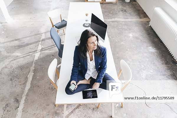 Nachdenkliche Geschäftsfrau sitzt auf dem Schreibtisch in einem Loft mit elektronischen Geräten.
