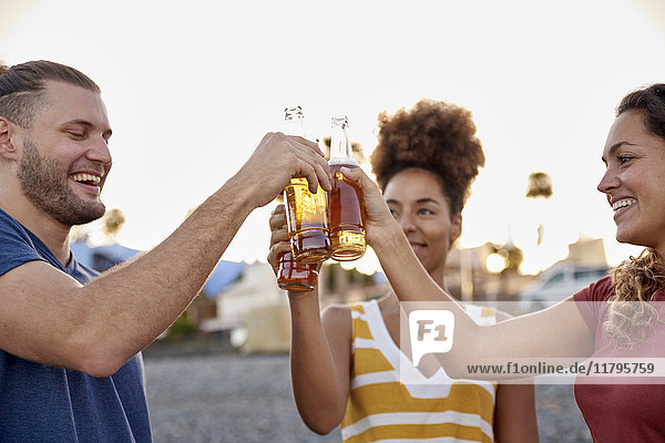 Drei Freunde stoßen mit Bierflaschen am Strand bei Sonnenuntergang an.