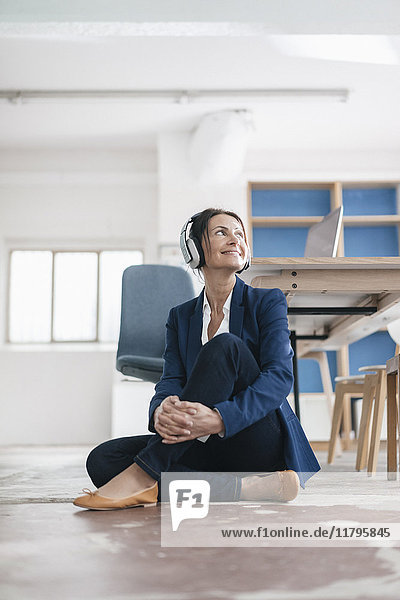 Lächelnde Geschäftsfrau sitzt auf dem Boden in einem Loft und hört Musik mit Kopfhörern.