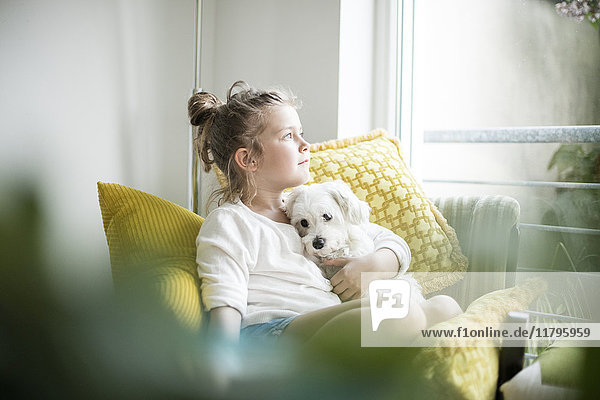 Kleines Mädchen sitzt auf einem Sessel zu Hause und kuschelt mit ihrem Hund.