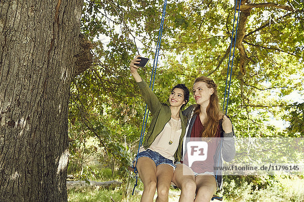 Zwei beste Freunde sitzen zusammen auf einer Schaukel und nehmen Selfie mit dem Smartphone.