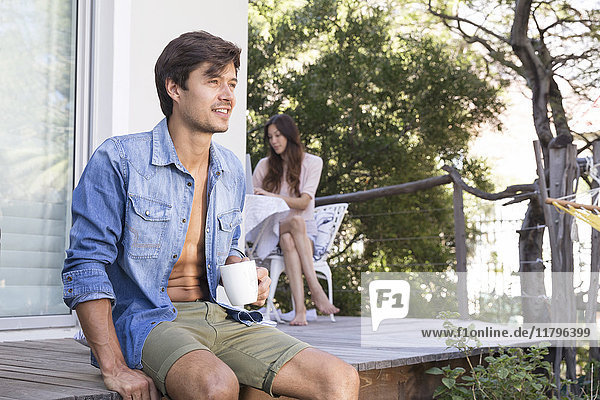 Man sitting on terrace with cup of coffee and woman in background
