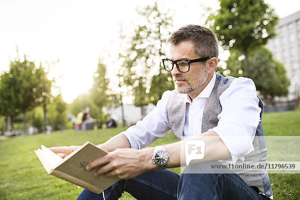 Confident mature businessman in the city park sitting on grass reading book