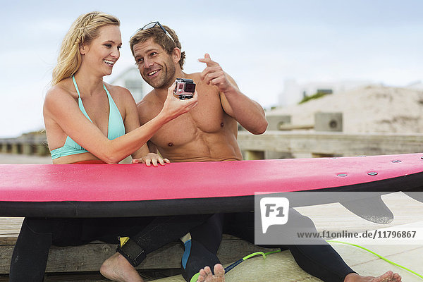 Glückliches Paar mit Surfbrett und Kamera am Strand