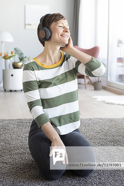 Porträt einer lächelnden Frau  die im Wohnzimmer auf dem Boden sitzt und mit Kopfhörern Musik hört.