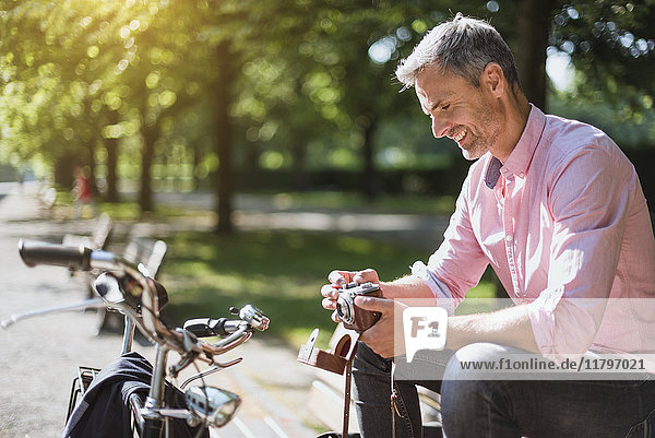 Lächelnder Mann mit Fahrrad  der auf einer Parkbank eine altmodische Kamera ansieht