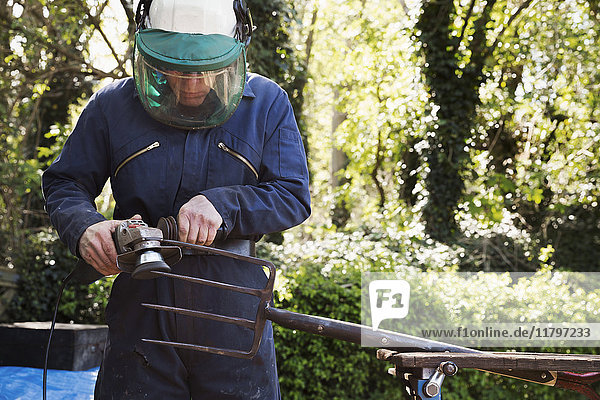 Man standing outdoors  wearing a face mask  working on a large metal garden fork or pitchfork with an angle grinder.