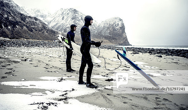 Two surfers wearing wetsuits and carrying surfboards standing on a beach with mountains behind.