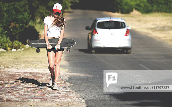 Junge Frau  die mit einem Skateboard neben einer Straße läuft.