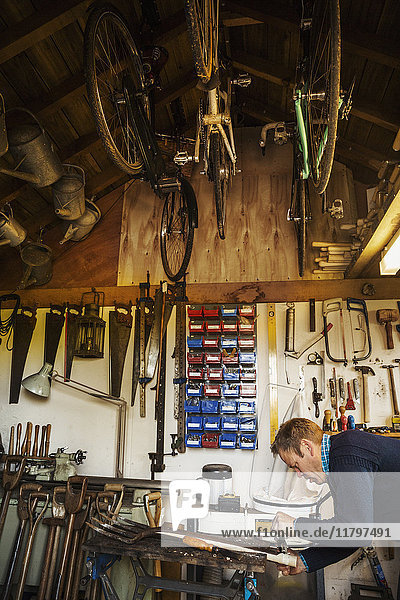 Ein Mann  der in einer Gartenwerkstatt steht und an einem alten Gartenplatz arbeitet  umgeben von Werkzeugen. Geräte an den Balken und Fahrräder  die von der Decke hängen.