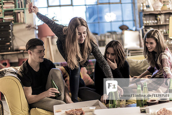 Drei junge Frauen und ein junger Mann sitzen lächelnd auf einem Sofa  Pizza und Bierflaschen auf dem Couchtisch.
