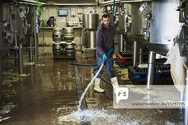 Mann  der in einer Brauerei arbeitet und den Boden mit einem Wasserschlauch reinigt.