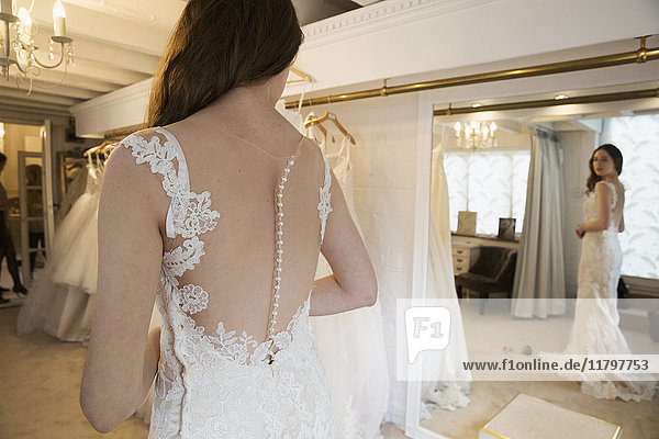 Eine junge Frau in einem weißen Hochzeitskleid in voller Länge  die ihr Spiegelbild im Spiegel in einer Brautboutique betrachtet.