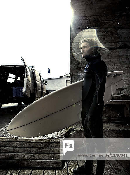 Ein Surfer  der einen Neoprenanzug trägt und ein Surfbrett trägt  steht an einem Holzgebäude und einem Lieferwagen.