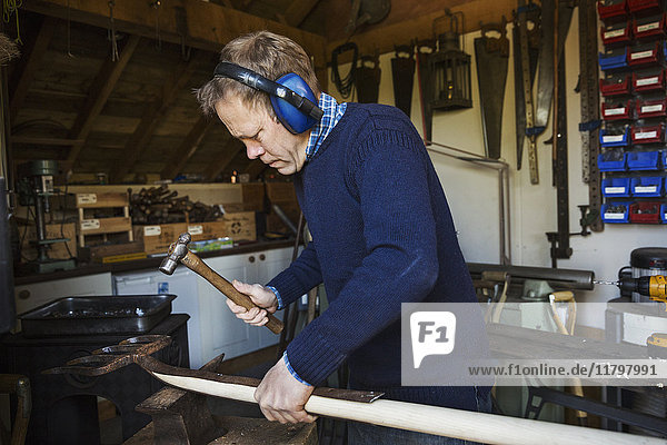 Mann steht in einer Werkstatt  trägt Gehörschutz  hält einen Hammer  arbeitet an einem Stück Holz.