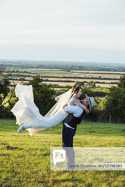 Neuvermählte stehen auf einer Graswiese mit Blick auf eine ländliche Umgebung  der Bräutigam umarmt und schwingt die Braut in die Luft.