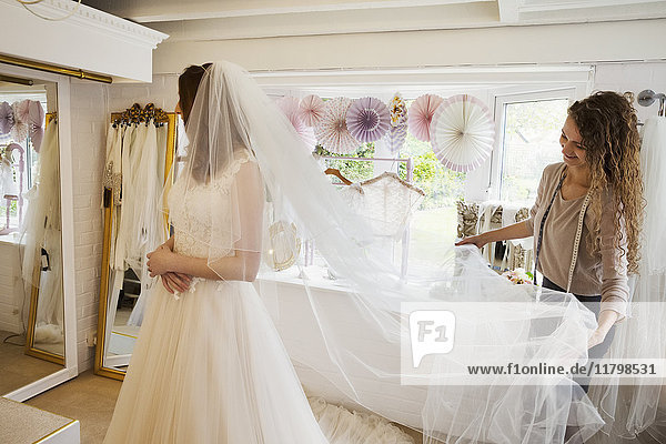 Eine junge Frau in einem weißen Hochzeitskleid in voller Länge und eine Assistentin oder Schneiderin  die ihren langen Netzschleier hochhält.