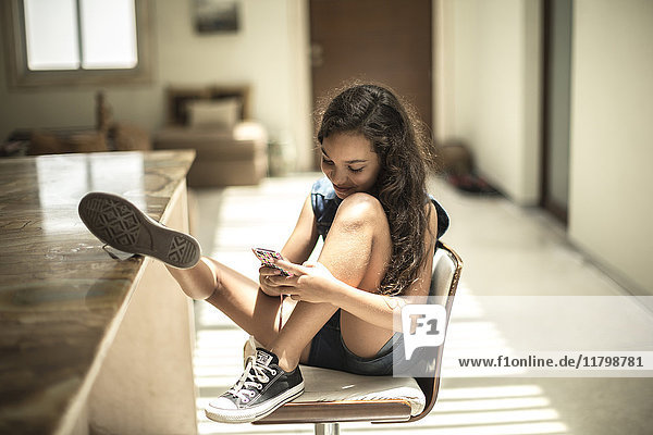 A girl sitting looking at a mobile phone screen with feet up on a kitchen counter.