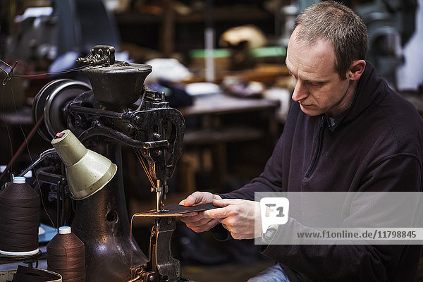 Man sitting at a large leather stitching machine in a shoemaker's workshop.