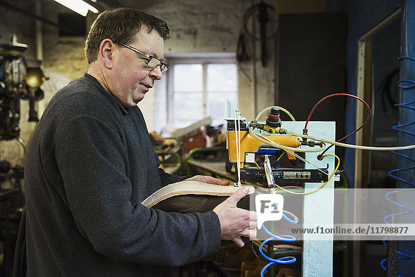 Man standing in a shoemaker's workshop  using a machine to sew a sole onto a shoe.
