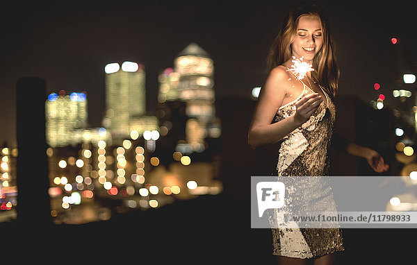 A young woman in a sequined dress dancing on a rooftop at night holding a party sparkler.