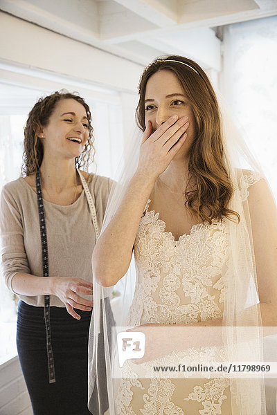 A young woman trying on a wedding dress  with lace overlay on the bodice and skirt  her hand covering her mouth  expressing surprise and delight. A dressmaker in the background.