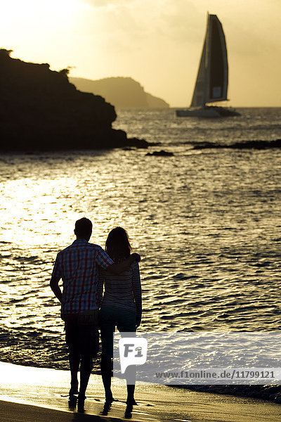 Rückansicht eines Paares  das bei Sonnenuntergang an einem Sandstrand am Meer  an Klippen und einem Segelboot in der Ferne steht.