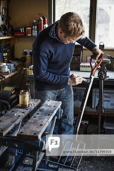 Man standing in a workshop oiling the handle of a metal garden fork with four tines.