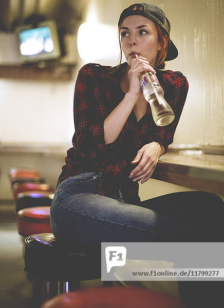 A woman sitting on a stool drinking from a glass bottle in a bar and looking over her shoulder.