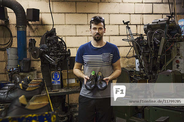 Man standing in a shoemaker's workshop  holding a pair of brown leather laced cycling shoes.