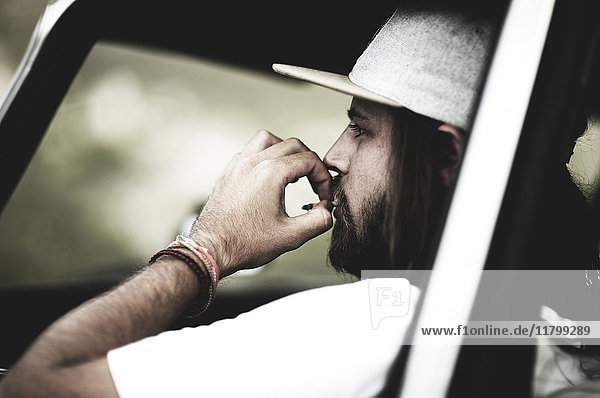 Bärtiger junger Mann mit Baseballmütze sitzt in einem Auto und raucht Zigaretten.