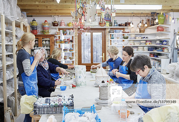 A group of people seated at a workbench in a pottery workshop  handbuilding clay objects. A woman with a cup of tea.