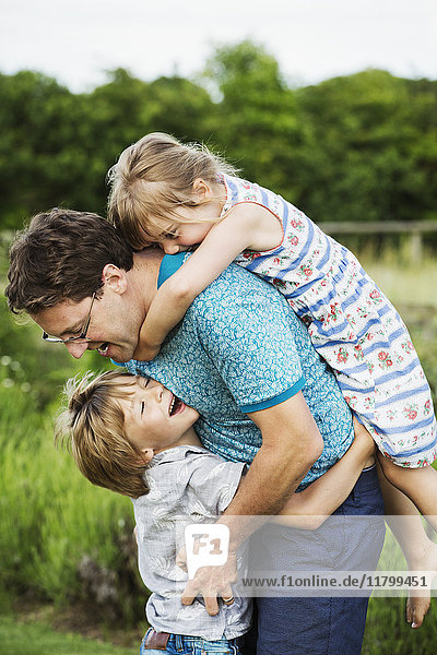 Ein Vater und zwei Kinder in einem Garten  ein Junge umarmt ihn um die Taille und ein Mädchen auf dem Rücken mit Armen um den Hals.