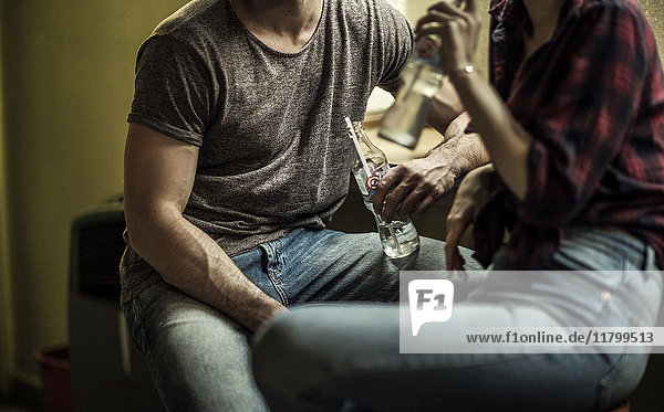 A man and a woman sitting and drinking from glass bottles on stools at a bar.