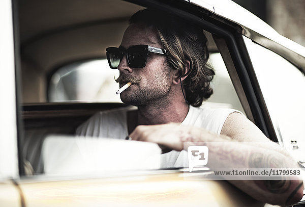 Mann mit Schnurrbart sitzt im Auto  trägt eine Sonnenbrille  raucht eine Zigarette  tätowierter Arm lehnt aus dem Fenster.