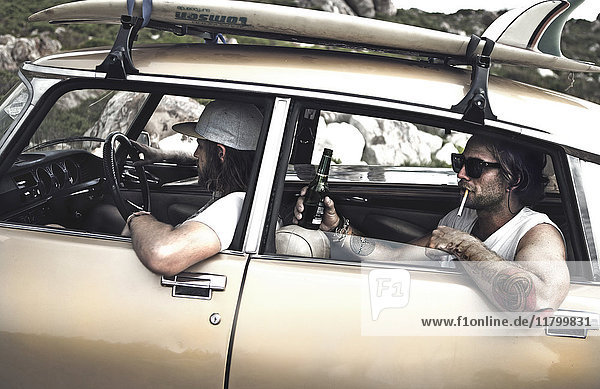 Two men in a vintage car with a surfboard on the roof  man on back-seat with cigarette in his mouth  holding a beer bottle  tattooed arm leaning out of window.