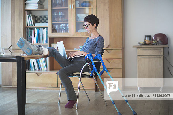 Woman resting her broken leg on table and using laptop on chair