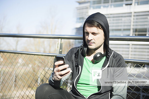 Mann in Sportkleidung mit Kopfhörern und Smartphone entspannt sich nach dem Training auf einer Brücke