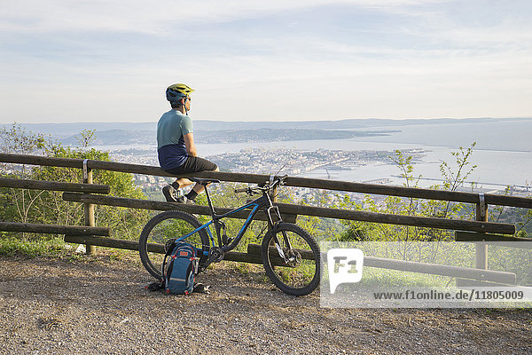 Mountainbiker sitzt auf einem Holzgeländer und schaut auf das Meer und die Stadt