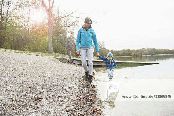 Mutter und Sohn gehen Hand in Hand vor einem Steg am Seeufer im Herbst spazieren
